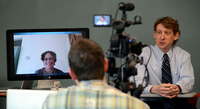 Cary Roseth at a desk, filmed during a video-call conversation with a colleague shown on a computer monitor